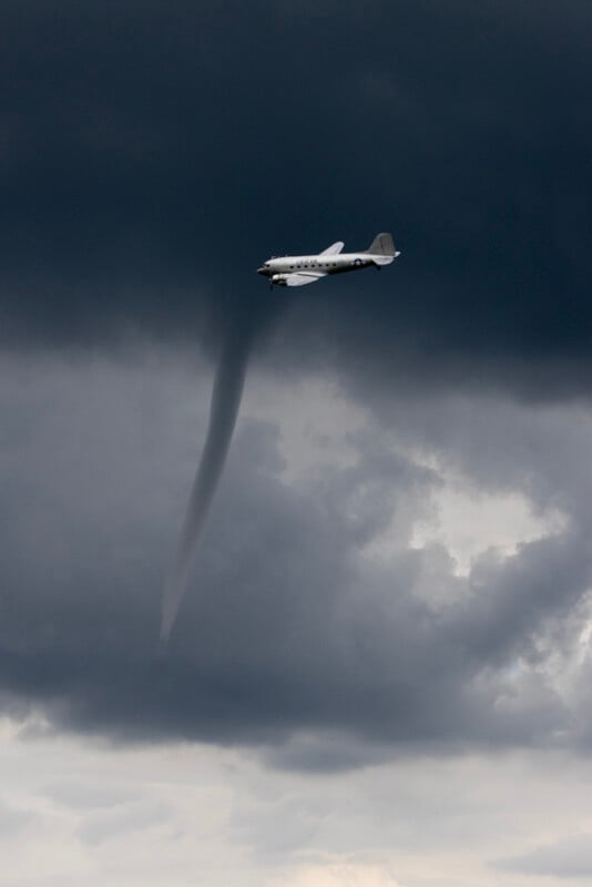 A vintage airplane flies near a dark tornado under stormy clouds, with the funnel extending from the sky toward the ground.