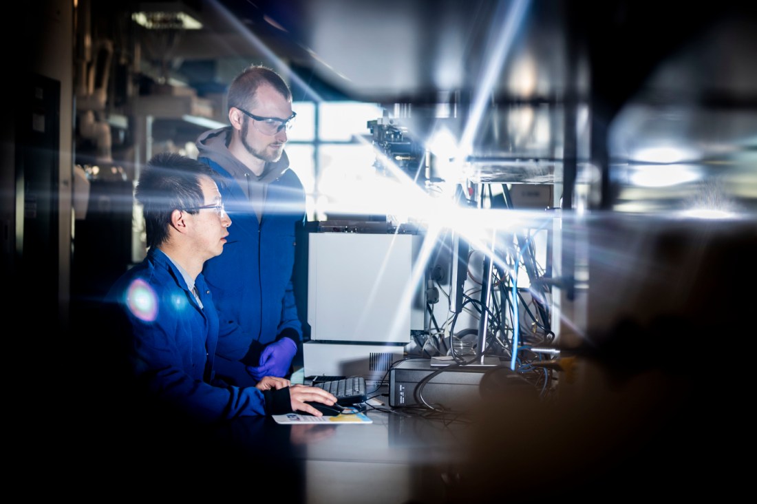 Two men wearing lab coats and goggles observe a computer in a laboratory.
