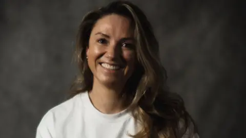 ONE LDN Picture of a woman smiling in a white t-shirt. She has brown hair and is in front of a grey backdrop.
