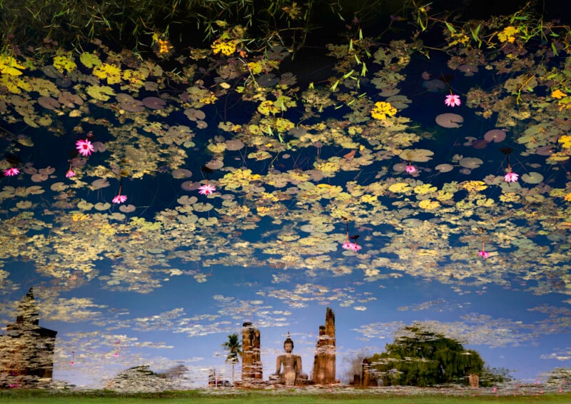 Lily pads and pink flowers float on water, reflecting a blue sky and the image of an ancient Buddha statue and temple ruins, creating a surreal, upside-down scene.