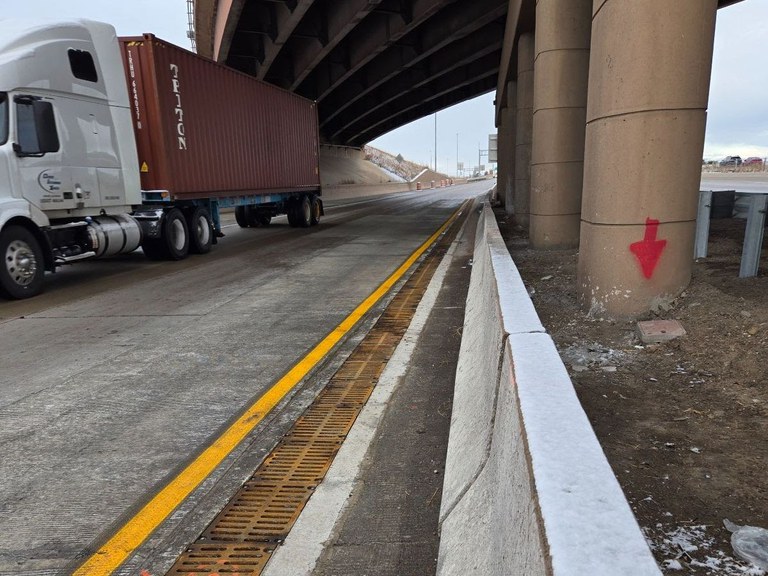 Drainage improvements along the edge of the roadway at I-70 off-ramp toward Airport Boulevard. A semi-truck and overpass are in the background.
