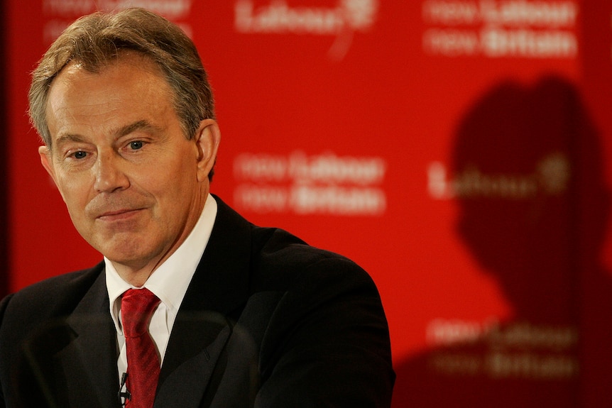 A man in a black suit with a red tie looks down 