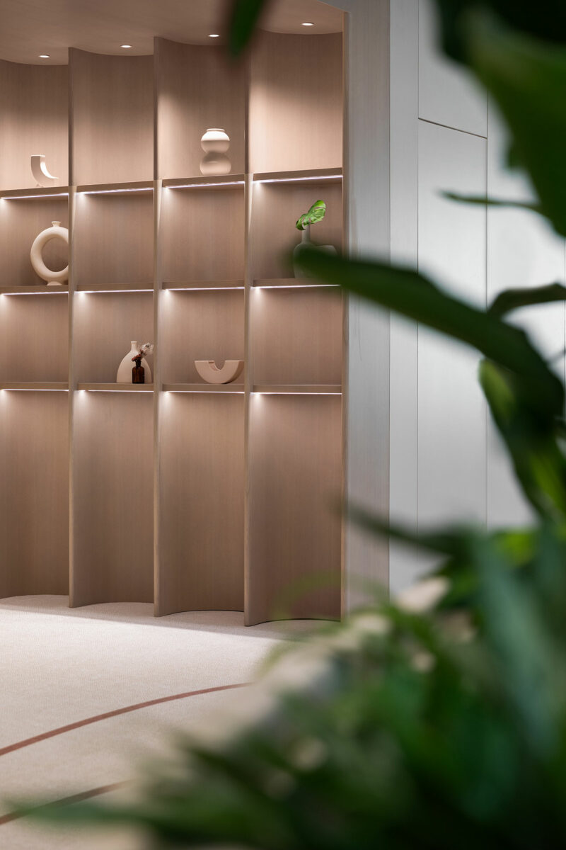 A modern wooden shelving unit with minimalist decor pieces is softly lit, with large green plants in the foreground.