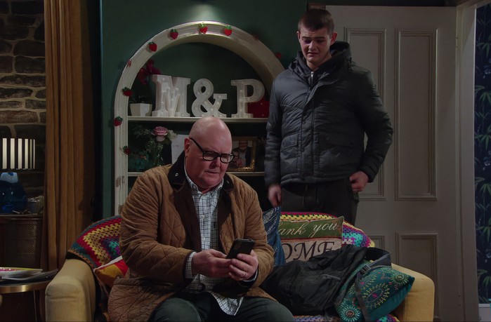 Dylan stands over Paddy, who is talking to Mandy on his phone, in Emmerdale