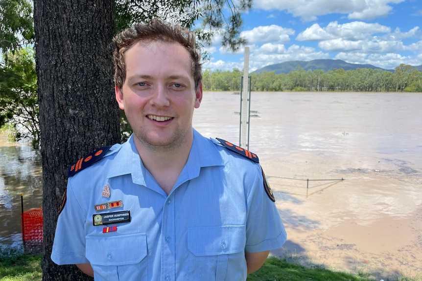 Young man in blue uniform, smiling at camera, flooding river behind, green grass