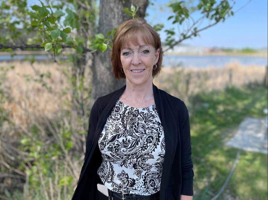 A woman outdoors in front of a tree and grassy area, smiling at the camera in a black jacket and patterned shirt.