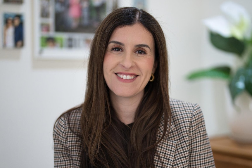 A smiling woman with long brown hair, wearing a light brown checked blazer, sitting in an office.