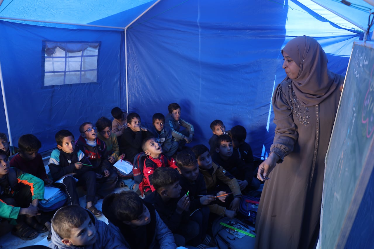 A woman in a beige dress and matching hijab writes on a chalkboard and looks out of her class of small children huddled inside a blue tent.