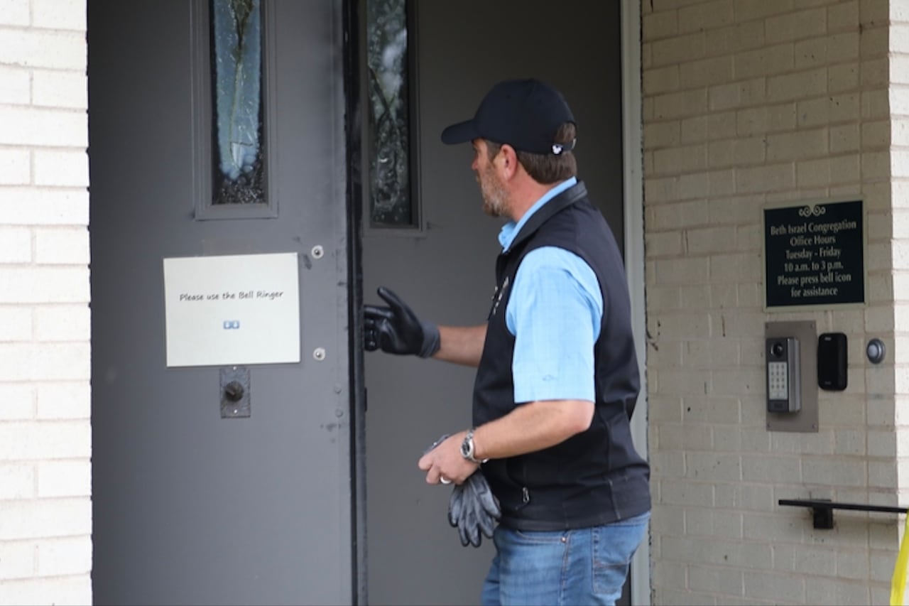 A man in a baseball cap, wearing gloves and a vest, is shown near a front door of a building. 