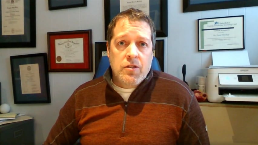 A man with short brown hair, wearing a sweater, sitting in an office, with several framed diplomas on the wall.