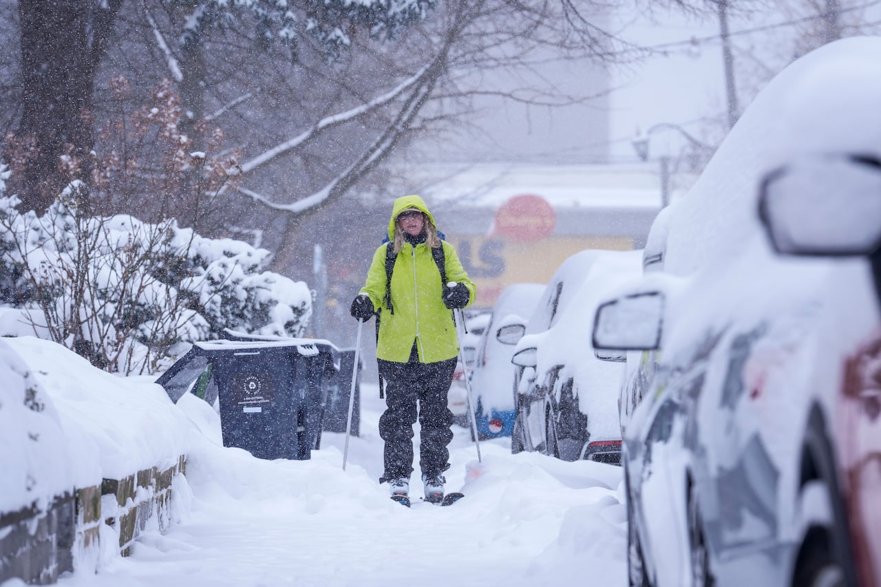 A woman cross-country skiing on a sidewalk next to parked cars during a snowy day.