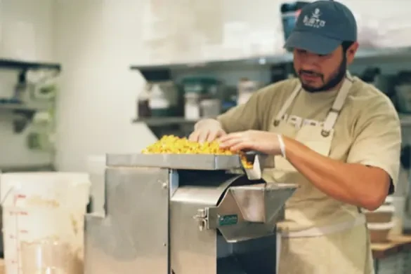 A man with medium skin tone, wearing a tan shirt, tan apron, and black baseball hat, grinds nixtamal in a kitchen restaurant.