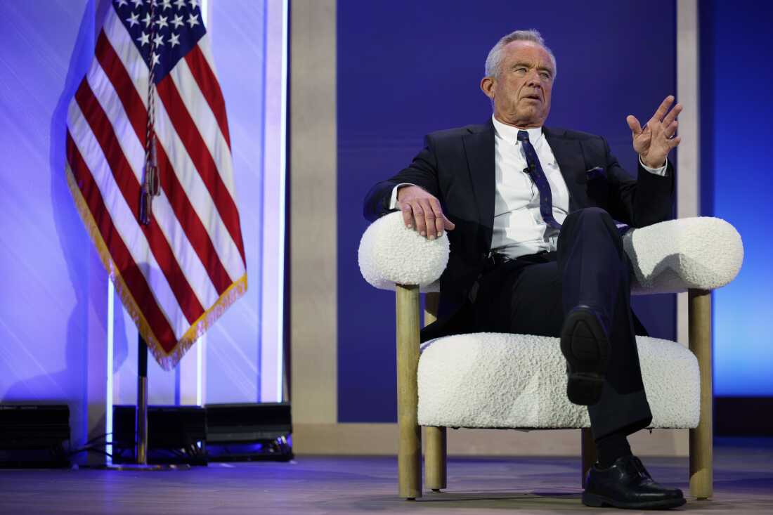 Secretary Kennedy is shown seated in a modern white armchair. An American flag is on his left. 