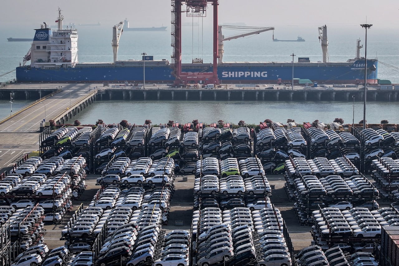Multi-level metal trailers stacked with cars are parked on a paved lot in front of a shipping port, with a big red crane and a large cargo vessel in the background. 