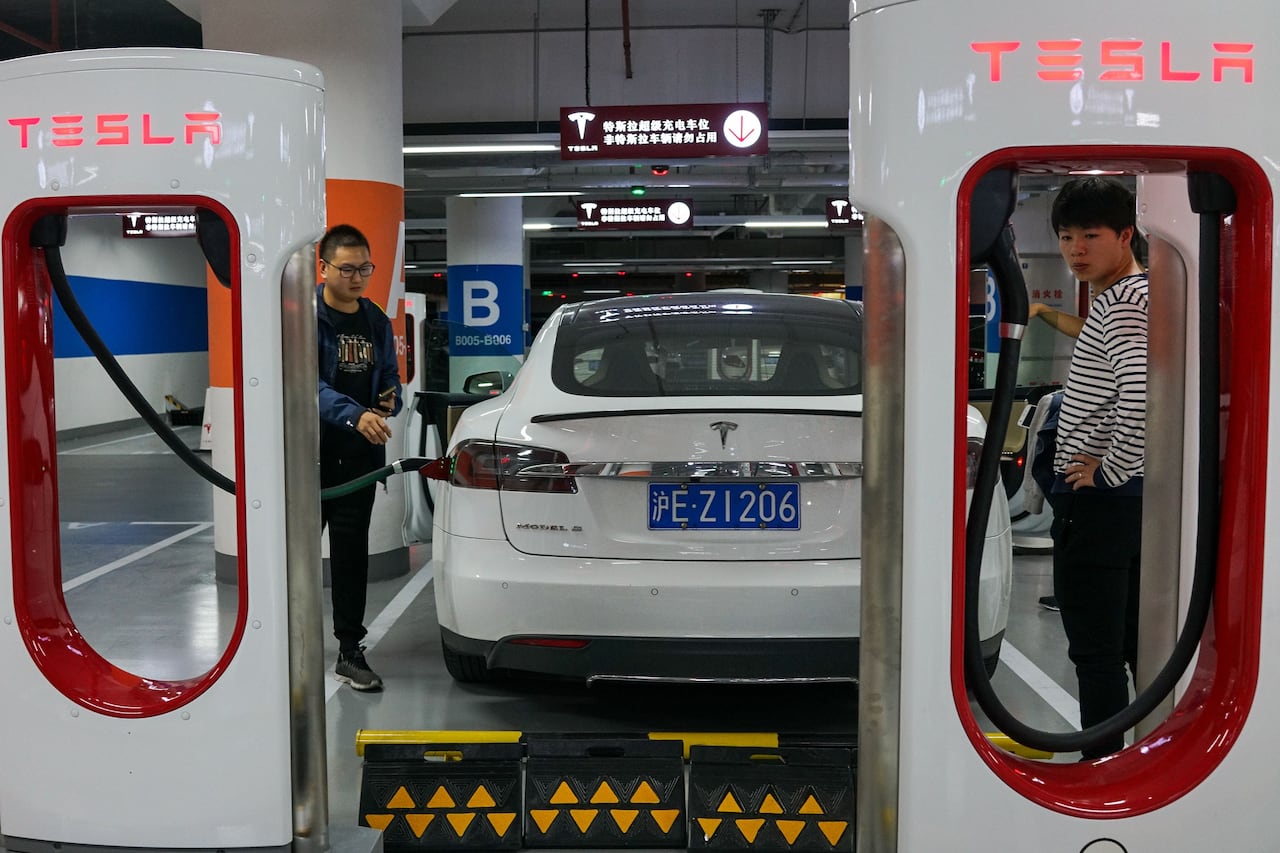 Two, tall white and red plastic EV charging stations with the word "Tesla" in red letters, at the top of each. In the background, A man on the left reaches for a charging cable affixed to a white car, while a woman on the right watches.