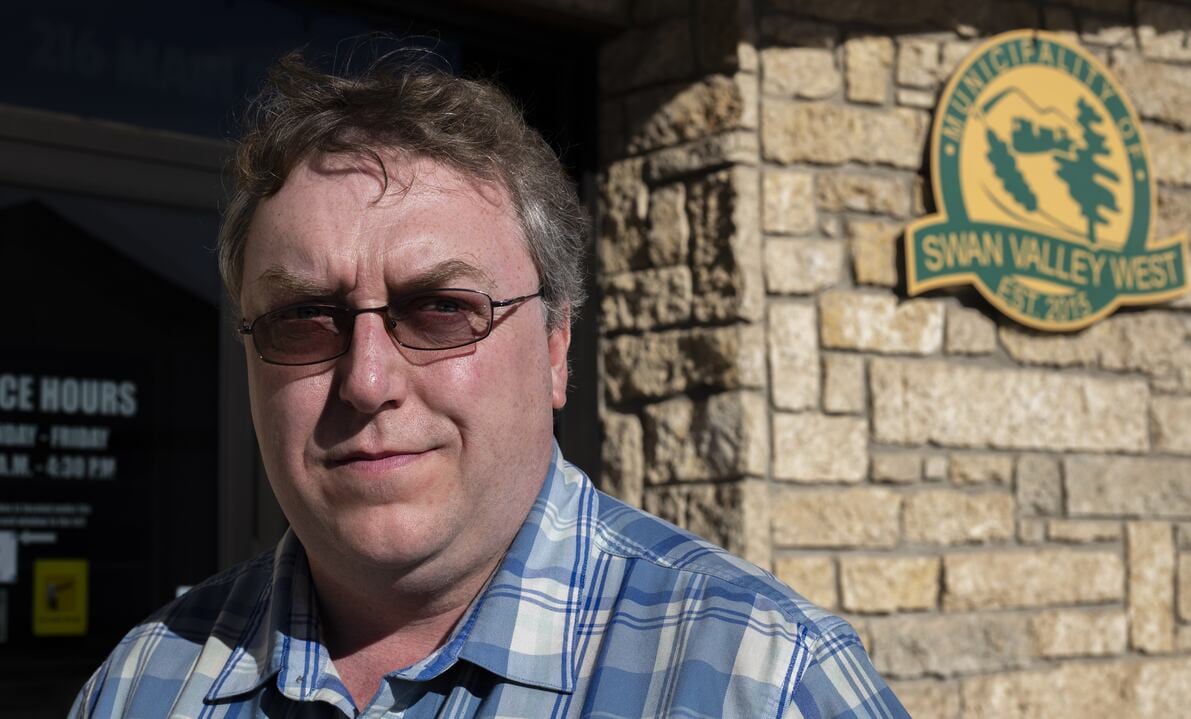 A man in a blue and white checkered shirt wearing glasses is looking into the camera. Behind his left shoulder is a sign that says Municipality of Swan Valley West.   