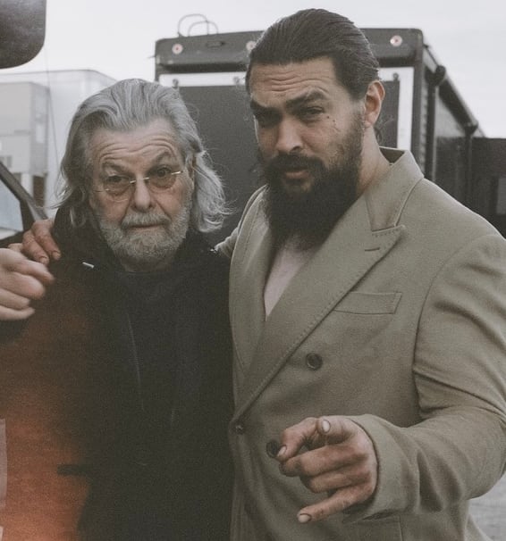 A man with a white beard stands next to a taller man with a brown beard, who is pointing at the camera. 