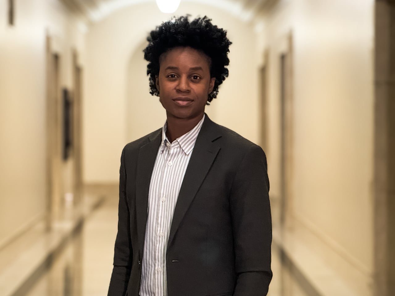 A person in a shirt and jacket, standing in a hallway, looks at the camera 