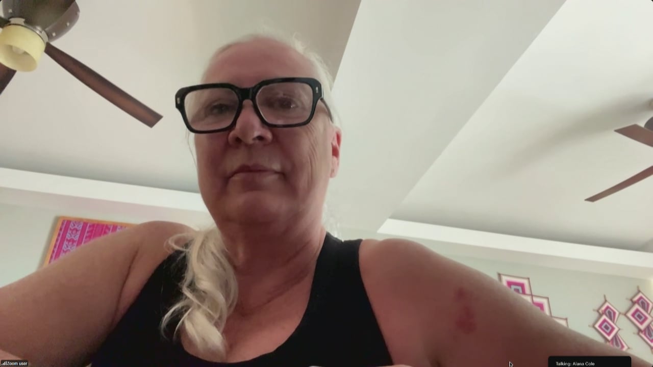 A woman with long grey hair wearing a black tank top looks into the camera.