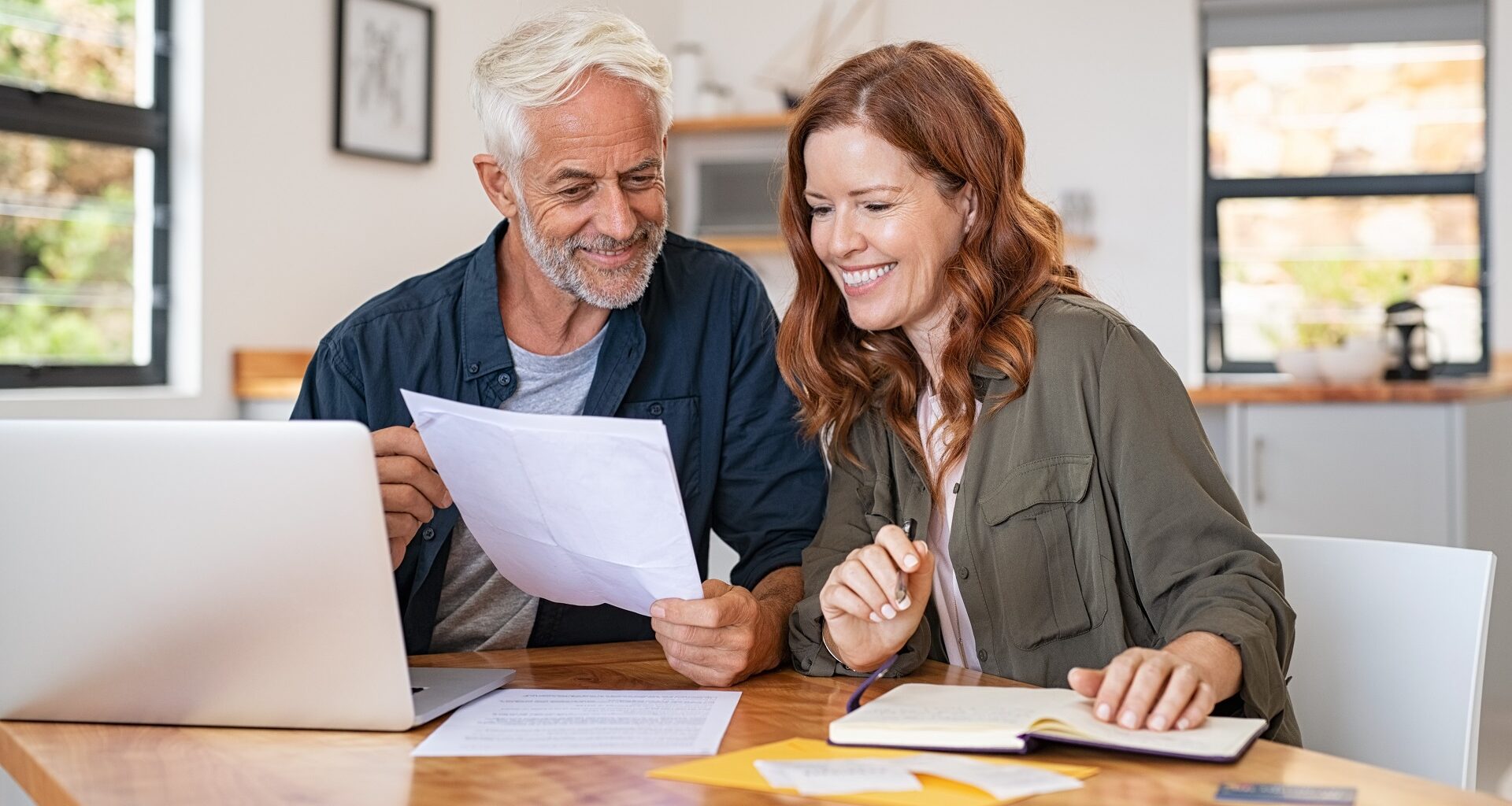 Mature couple sitting at a table, holding papers and working on their finances on a laptop in their house