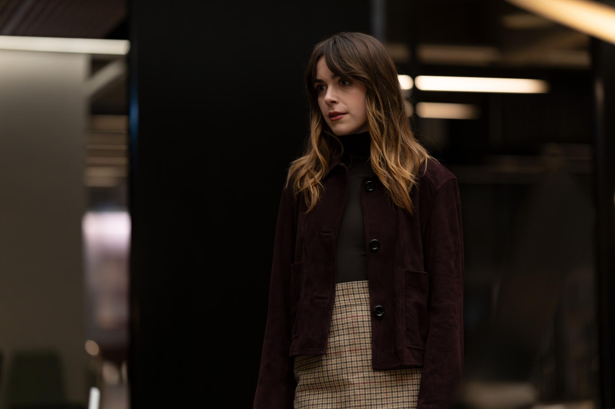 A woman with long brown hair in a black turtleneck, maroon coat and plaid skirt.