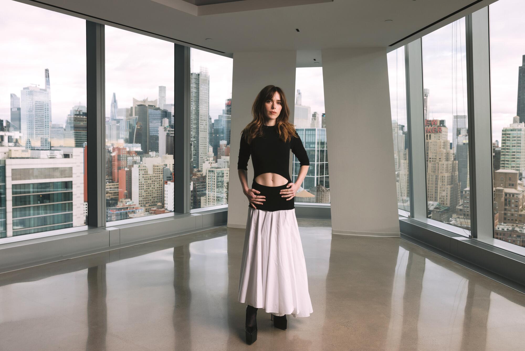 A woman in a black top and white skirt, baring her midriff, stands near a windowed corner overlooking the New York skyline.