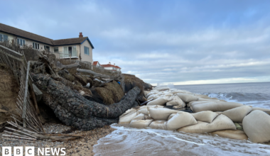 A home on the edge of an eroding cliff in Thorpeness. There is a big cluster of geobag sea defences on the beach in front of it. The tide is washing in.