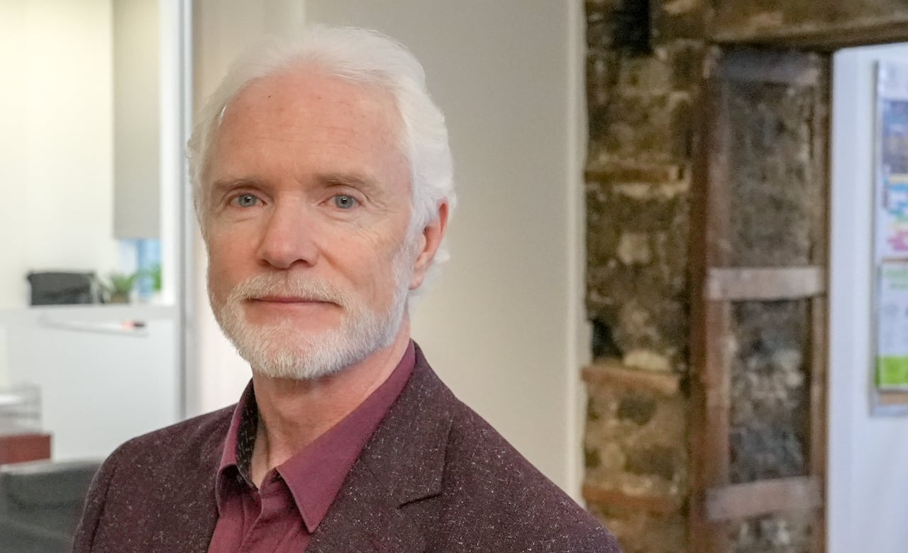 A man with a white beard, white hair and a burgundy suit stands in an entrance hallway.