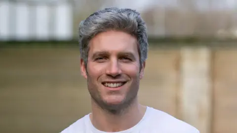 Michael James Close up of Dafydd smiling looking at the camera. He is standing in front of a brick wall but the background is blurred. He wears a white t-shirt and has grey hair.