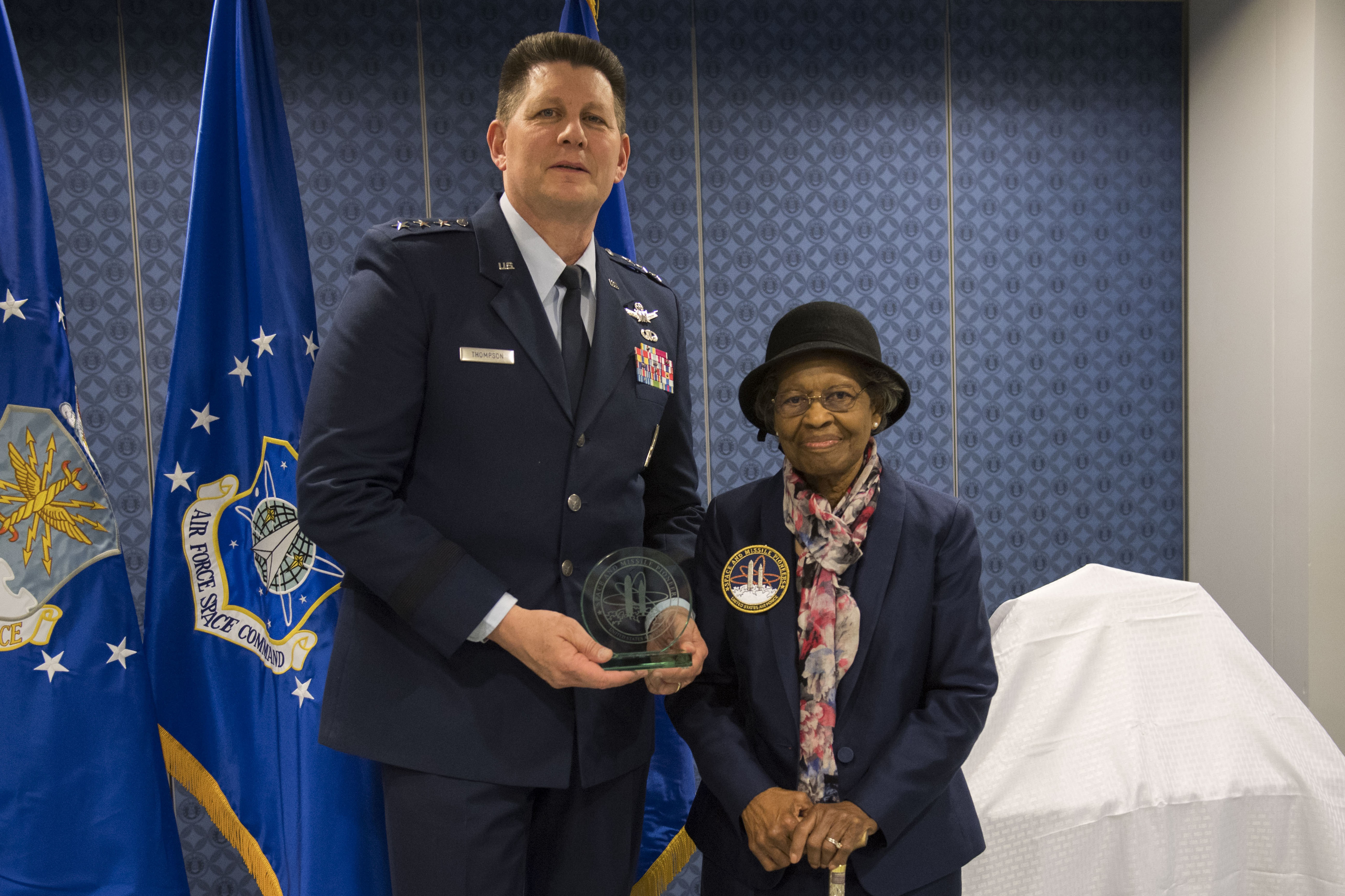 A uniformed man holds an award next to an elderly woman in a hat, standing before blue Air Force flags in a formal setting.