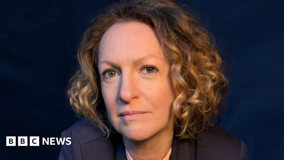 A professional headshot of Victoria Woodall with chin length golden curly hair wearing a navy suit against a navy backdrop
