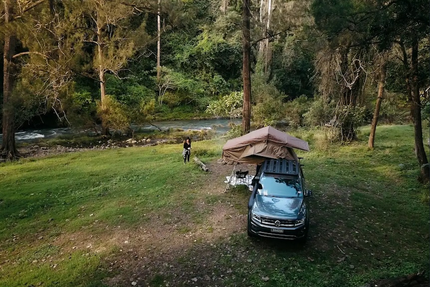 A car with a camping trailer on a grassy area with trees and a creek in the background.