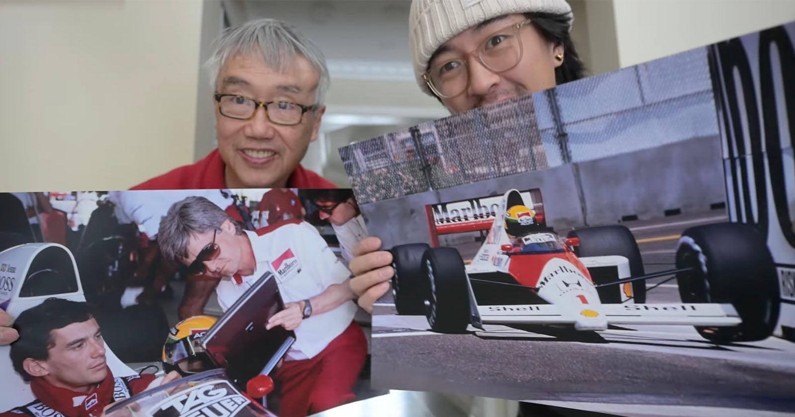 Two men smile at the camera while holding large photos of Formula 1 scenes, including a driver signing an autograph and a racing car on the track.