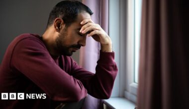 Man in maroon jumper with his head in his hands, sat by his window. He has dark hair and dark facial hair.