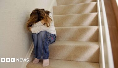 A generic picture of a girl sitting on a carpeted staircase in a home with their head in their hands. The girl is wearing jeans and white sweatshirt, she has long blonde hair but her face is obscured by her posture.