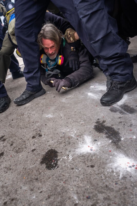 A man lies on the ground, partially restrained by several police officers in dark uniforms. The man appears distressed, gripping an object, while white powder is scattered on the pavement nearby.
