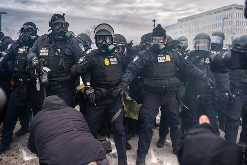 A line of police officers in riot gear and gas masks stand together during a tense outdoor protest, with some officers reaching out to restrain people in front of them. The sky is overcast and there is a sense of urgency.
