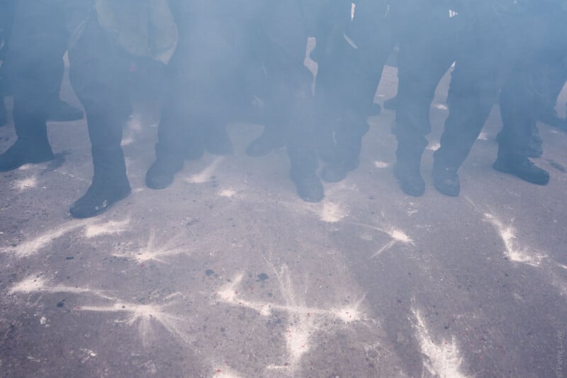 A group of people standing closely together, mostly visible from the knees down, surrounded by a cloud of smoke or tear gas, with white markings and powder scattered on the asphalt ground.