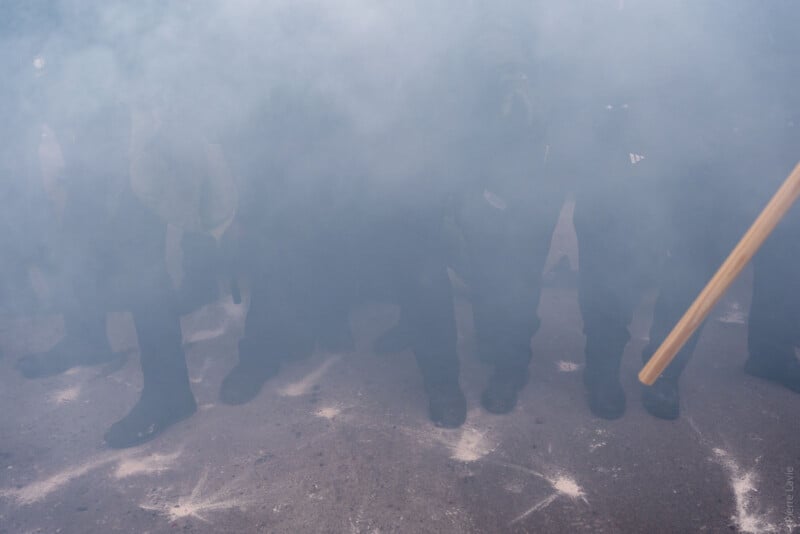 A group of people, mostly obscured by thick smoke or tear gas, stand close together on a dusty ground with visible white powder marks. Only their legs and feet are clearly visible. A wooden stick appears on the right.