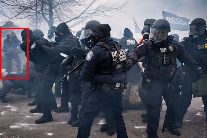 A line of police officers in riot gear face protesters amid smoke. One officer holds out an arm, and several flags, including one reading "RESIST," are visible in the background.