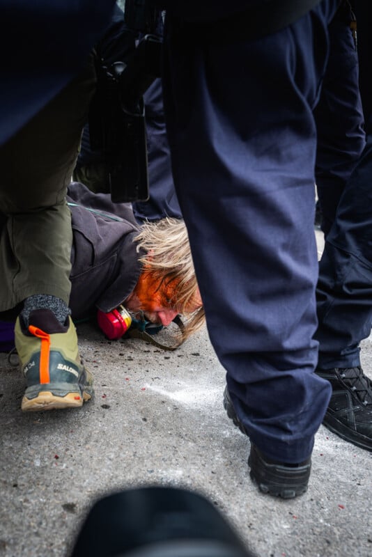 A person with a gas mask is lying on the ground, face partially covered in an orange substance, surrounded by the legs of several people, some in dark uniforms, suggesting a tense or chaotic situation.