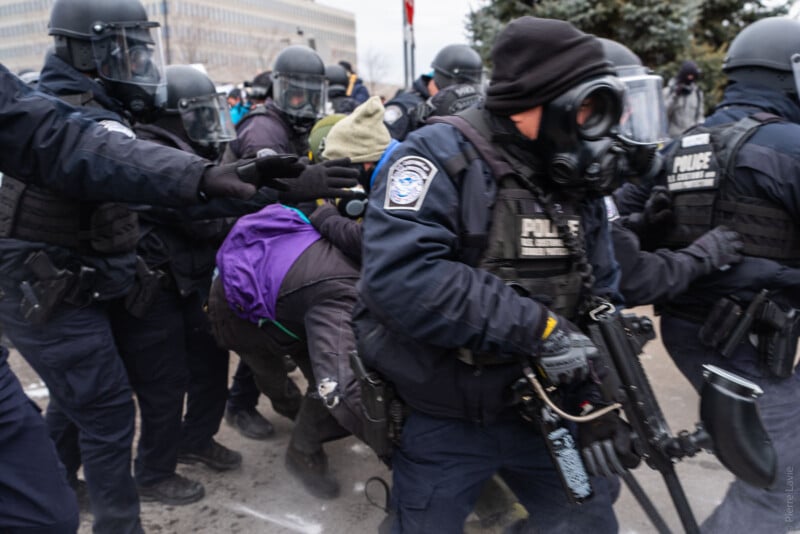 Police officers in riot gear detain a protester during a chaotic scene outdoors. Officers wear masks and helmets; one carries a firearm, while others reach toward the protester amid a crowd and tense atmosphere.