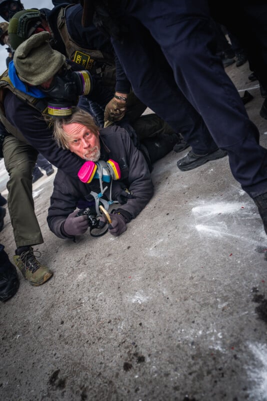 A person wearing a gas mask is pinned face-down on the ground by law enforcement officers, hands zip-tied, surrounded by people in dark clothing and boots on a concrete surface.