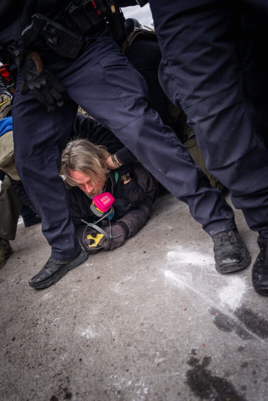 A person wearing a gas mask is on the ground being restrained by police officers in dark uniforms. The scene appears tense, with the person holding a pink respirator while surrounded by officers’ legs on a concrete surface.