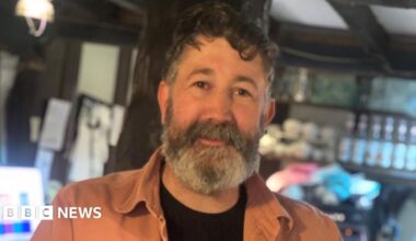 A middle-aged man with curly black hair and a grey beard. He is looking into the camera and is wearing a black t-shirt with orange shirt over the top. He is stood in a room with bottles and cups on shelving behind him, with a low beamed ceiling.