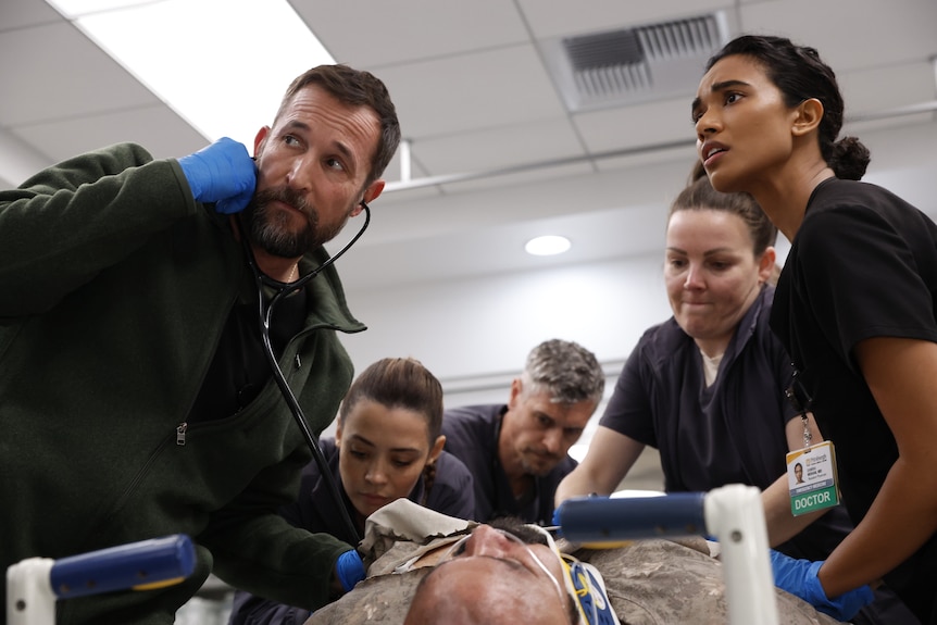 A TV still of Noah Wyle, 54, stethoscope in his ears, crowding, with Supriya Ganesh, 28, and more, over a person on a gurney.