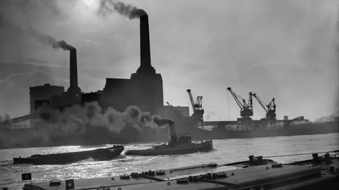 Fox Photos A black and white picture of Battersea power station in 1937 with billowing smoke coming from the towers