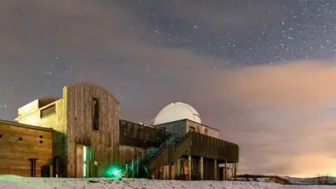 Steven Tsang A wooden-fronted observatory building on snowy ground underneath a very starry sky