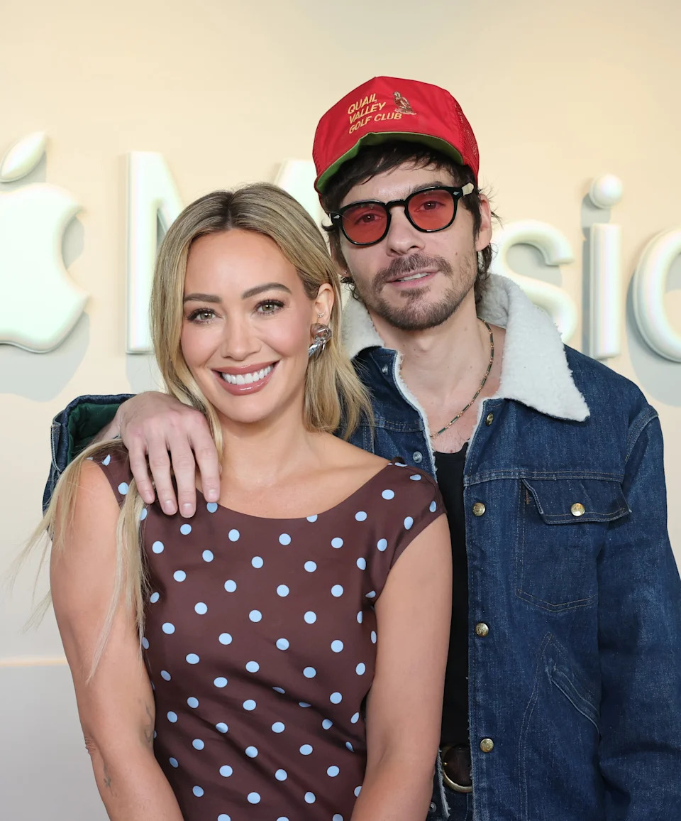 Two people pose in front of an Apple Music logo; one wears a polka dot dress, the other a denim jacket and red cap