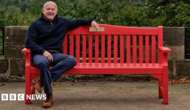 Dean Dyble sits on a bright red wooden red bench. He is an older man, with a bald head and a blue jumper, blue jeans and brown boots. In the background, trees and a stone wall can be seen.